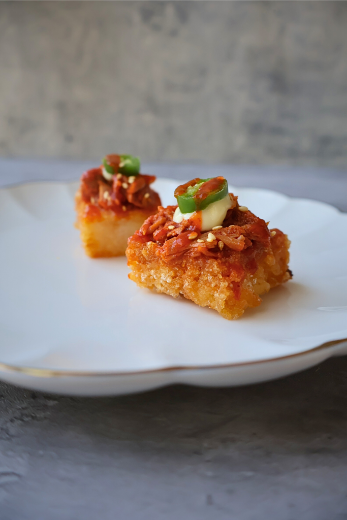 Close-up of two pieces of crispy sushi rice topped with spicy tuna, sliced jalapeños, and green onions on a white plate.