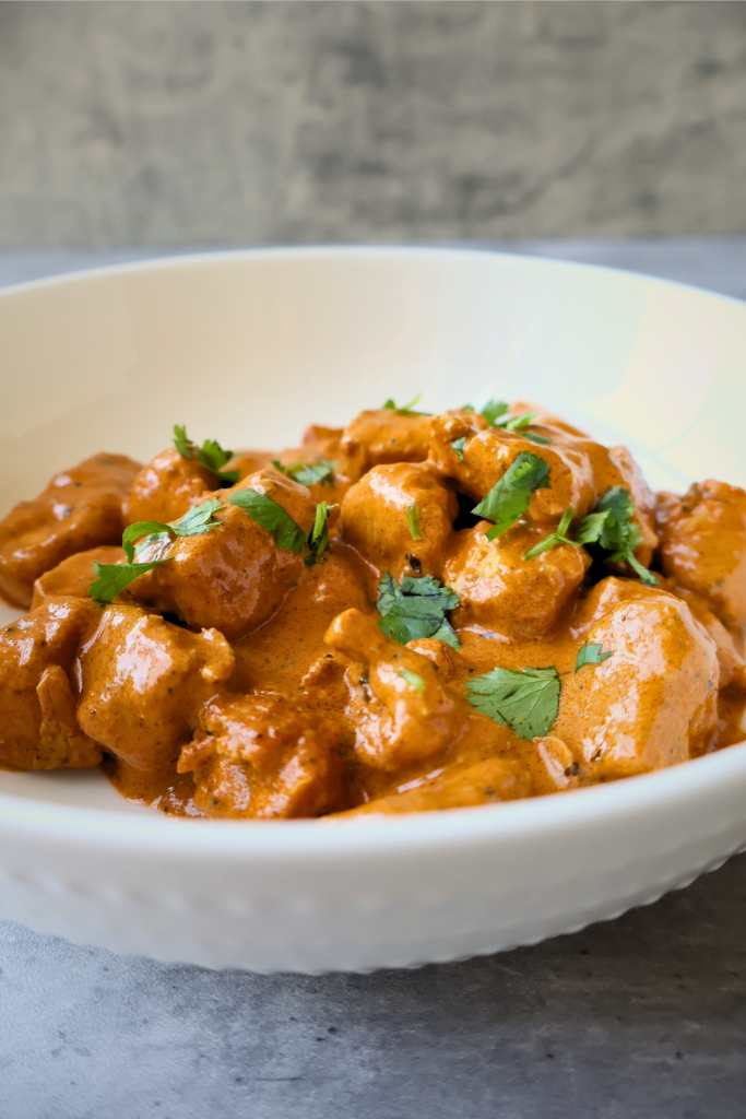 Creamy butter chicken served in a white bowl, featuring tender chicken pieces coated in a rich tomato and butter curry sauce, garnished with fresh cilantro.