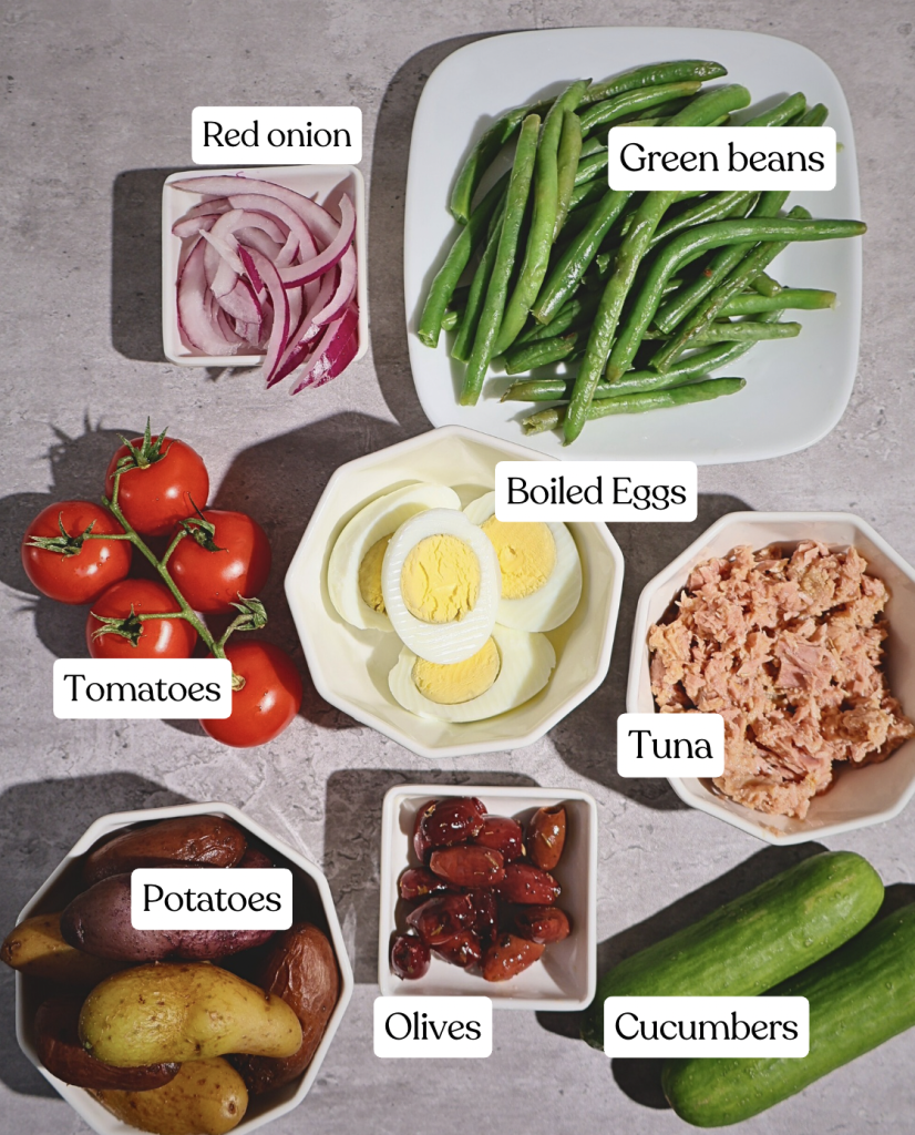 Ingredients for a Niçoise-style tuna salad arranged in small bowls, including tuna, boiled eggs, green beans, tomatoes, cucumbers, baby potatoes, red onion, and olives.