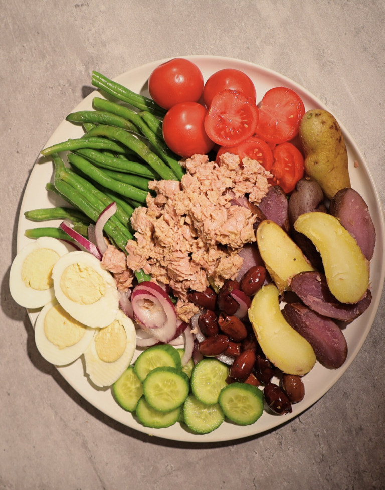 Niçoise-inspired salad with tuna, boiled eggs, baby potatoes, green beans, tomatoes, cucumbers, red onion, and olives arranged on a white plate.