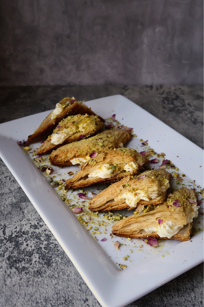 Traditional Middle Eastern warbat pastries filled with creamy ashta, arranged on a serving platter and topped with crushed pistachios and dried rose petals.