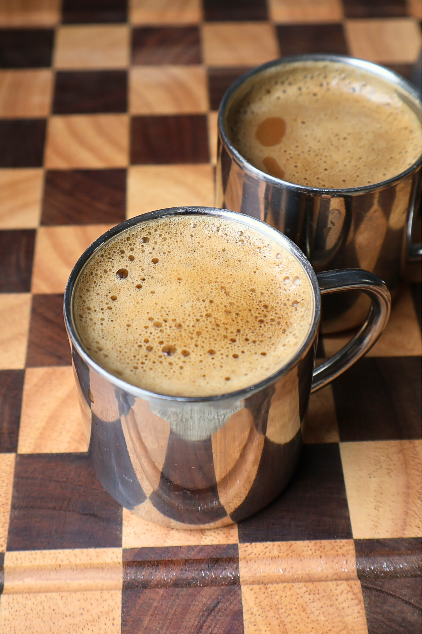 Two stainless steel mugs filled with freshly brewed coffee topped with rich crema, served on a wooden checkerboard board.