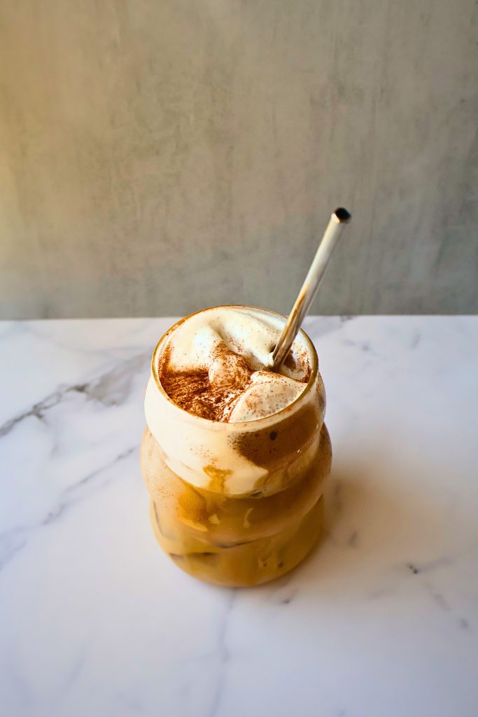 Glass of creamy iced coffee topped with frothy foam and a sprinkle of cinnamon, served with a reusable metal straw on a marble countertop.
