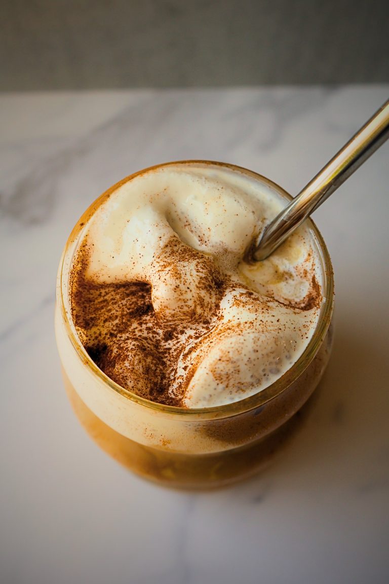 Creamy whipped coffee topped with frothy foam and a dusting of cinnamon, served in a glass with a metal straw.