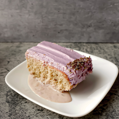 Slice of homemade tres leches cake topped with purple whipped frosting and colorful sprinkles, served on a white plate.