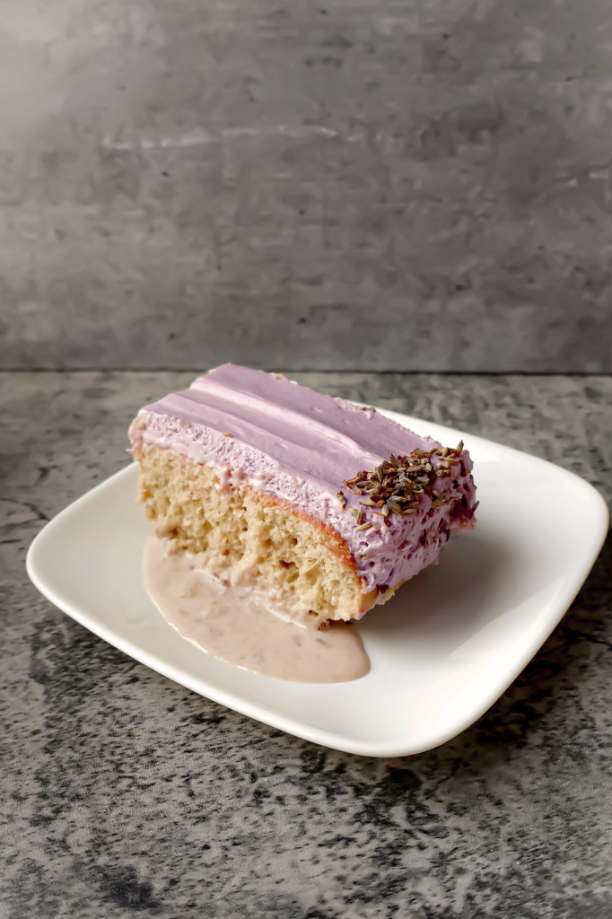 Slice of homemade tres leches cake topped with purple whipped frosting and colorful sprinkles, served on a white plate.