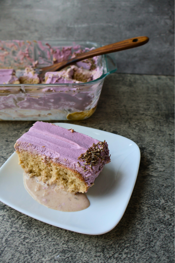 Slice of homemade tres leches cake with purple whipped frosting served on a white plate, with the baking dish in the background.