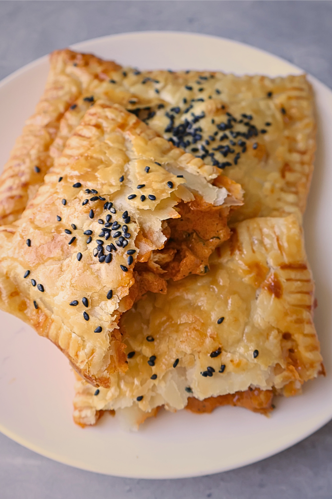 Golden, flaky phyllo pastry pockets filled with creamy butter chicken and topped with black sesame seeds, served on a white plate.