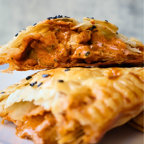 Close-up of a golden, flaky puff pastry filled with creamy butter chicken, topped with black sesame seeds, and served on a white plate against a light gray background.