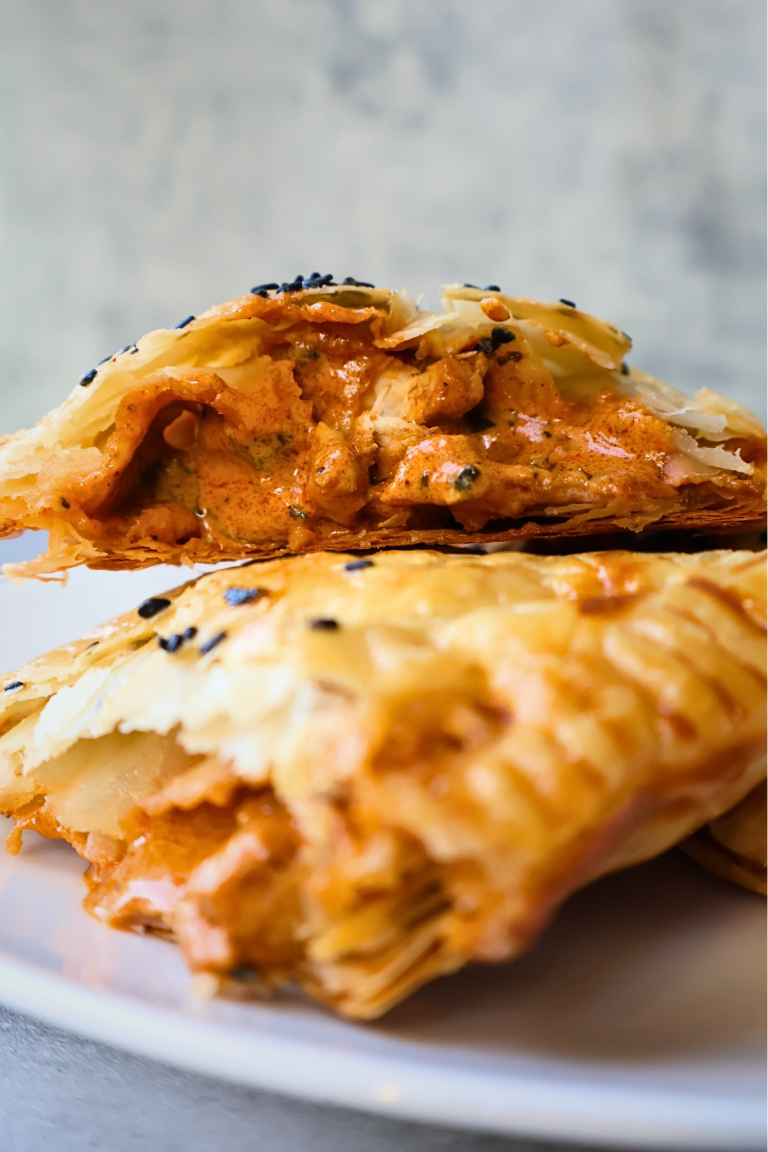 Close-up of a golden, flaky puff pastry filled with creamy butter chicken, topped with black sesame seeds, and served on a white plate against a light gray background.