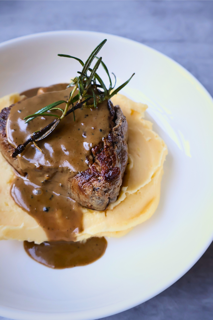 Pan-seared filet mignon served over creamy French aligot with a rich peppercorn sauce and fresh rosemary garnish.