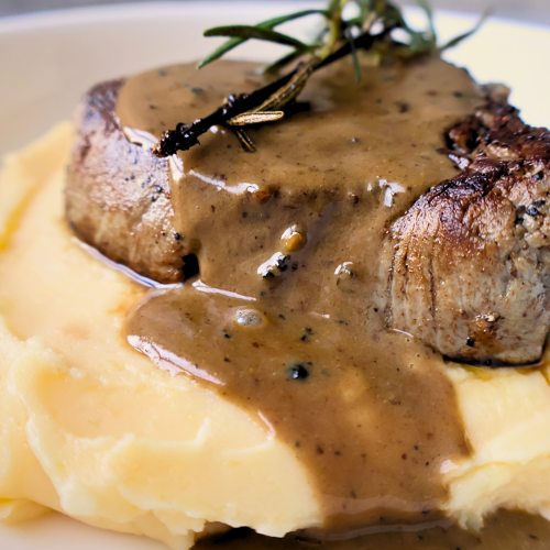 Close-up of filet mignon topped with peppercorn sauce and rosemary sprigs, served over creamy aligot mashed potatoes on a white plate.