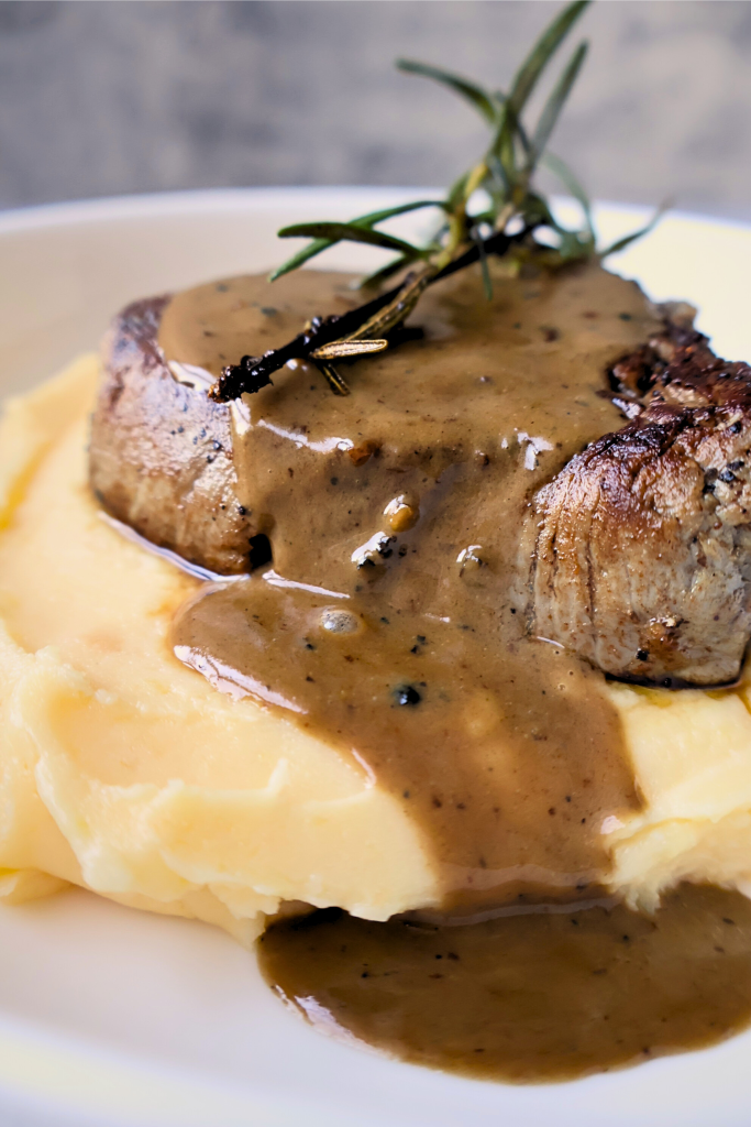 Close-up of filet mignon topped with peppercorn sauce and rosemary sprigs, served over creamy aligot mashed potatoes on a white plate.