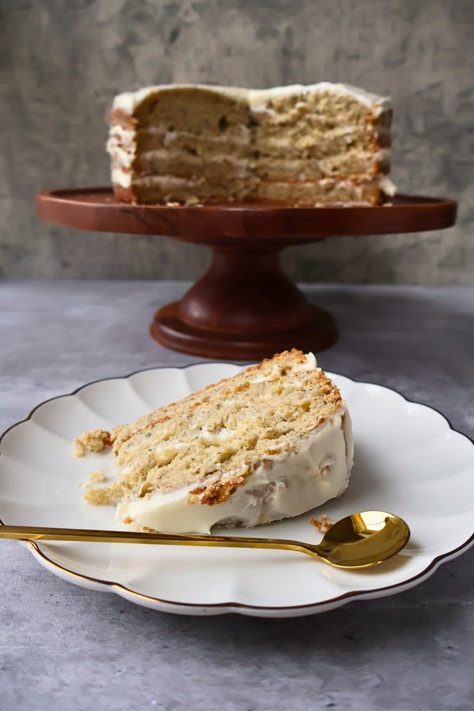 Slice of homemade banana cake with cream cheese frosting served on a dessert plate, with the remaining layered cake displayed on a wooden cake stand in the background.