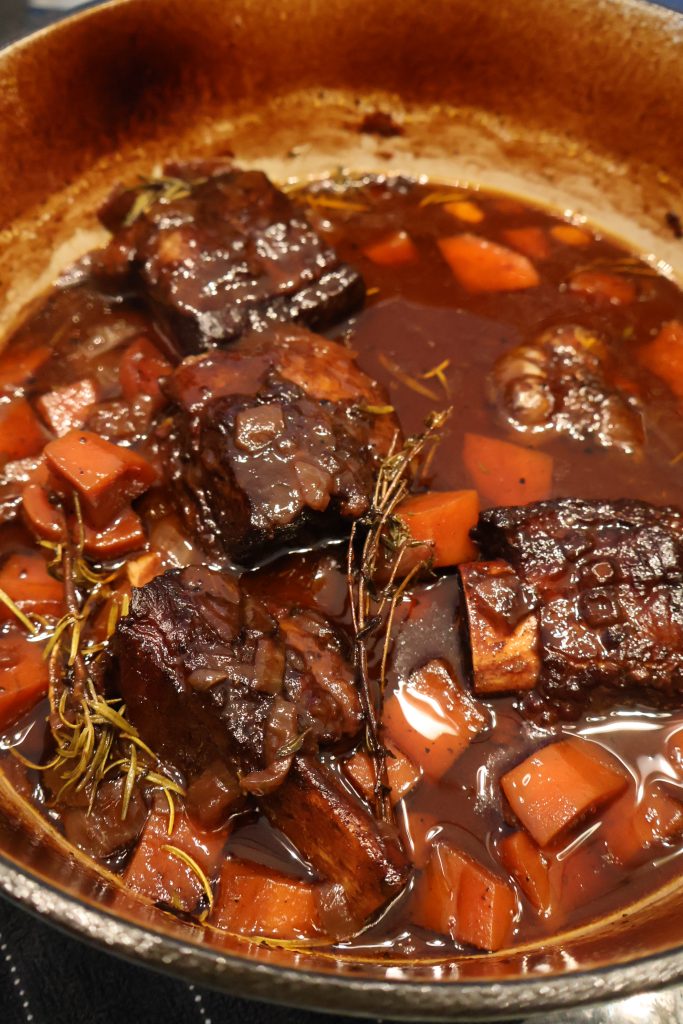 Dutch oven with slow-braising beef short ribs, carrots, herbs, and rich red wine sauce cooking until tender.