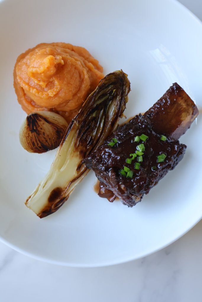 Braised beef short ribs plated with creamy mashed sweet potatoes, roasted baby onions, and charred leeks.