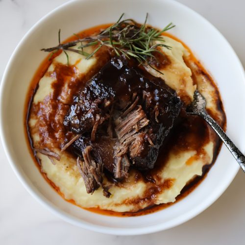 Maple and red wine braised short ribs over mashed potatoes, glazed in glossy sauce with rosemary garnish.