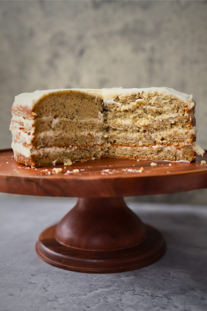 Slice of homemade banana cake with cream cheese frosting displayed on a wooden cake stand, showing its soft, moist layers.