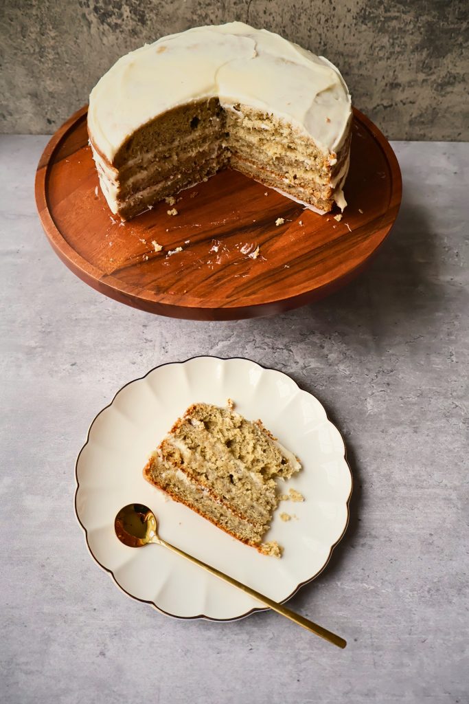 Whole homemade banana cake with cream cheese frosting on a wooden cake stand alongside a sliced piece served on a dessert plate.
