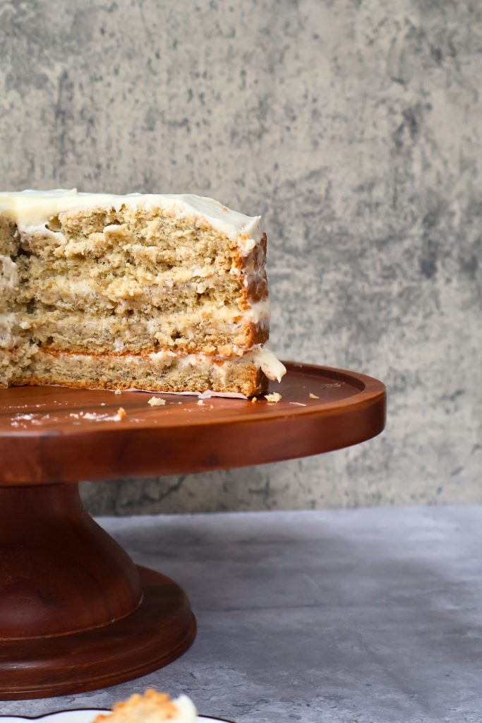 Homemade banana cake with cream cheese frosting displayed on a wooden cake stand against a neutral gray background.