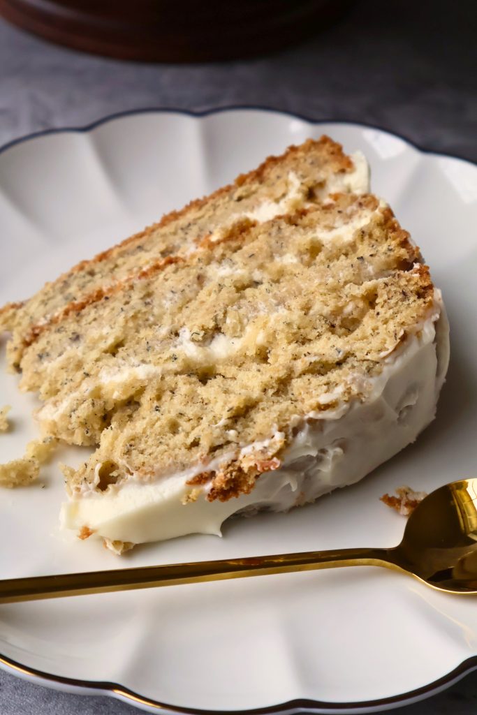 Close-up of a slice of moist banana layer cake with cream cheese frosting on a white scalloped plate beside a gold spoon.