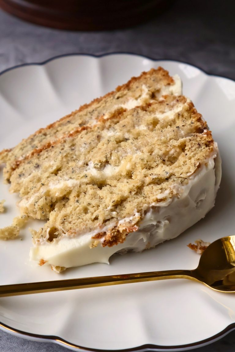 Close-up of a slice of moist banana layer cake with cream cheese frosting on a white scalloped plate beside a gold spoon.