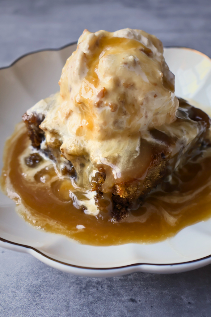 Sticky toffee pudding topped with vanilla ice cream and rich homemade toffee sauce served on a white dessert plate.