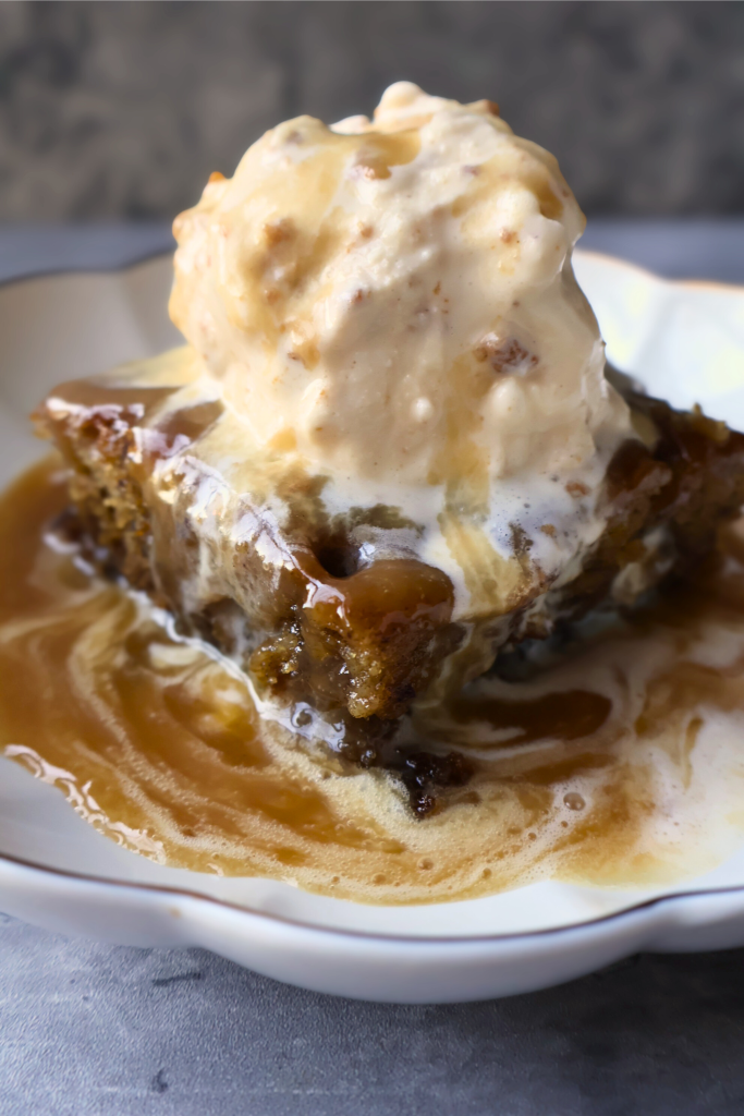 Sticky toffee pudding topped with vanilla ice cream and warm homemade toffee sauce in a white dessert bowl.