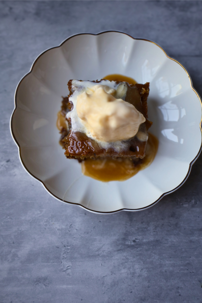 Overhead view of homemade sticky toffee pudding topped with vanilla ice cream and rich toffee sauce on a white dessert plate.