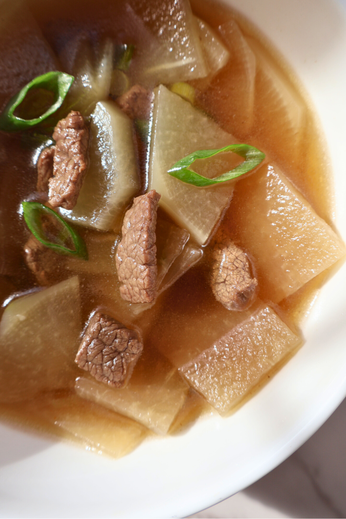 Close-up of Korean mu guk with beef chunks and soft radish slices in a clear broth, topped with fresh green onions