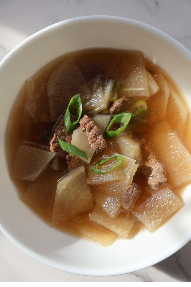 Overhead view of Korean mu guk in a white bowl with beef, radish slices, and green onions in a clear broth