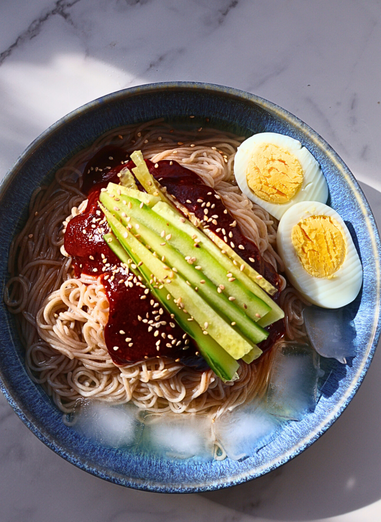 Korean bibim naengmyeon with spicy gochujang sauce, cucumber slices, sesame seeds, and boiled egg in a blue bowl