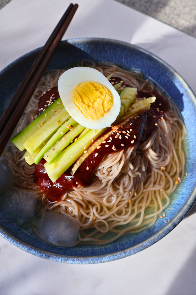 Top view of Korean bibim naengmyeon with spicy gochujang sauce, cucumber sticks, sesame seeds, and boiled egg in a blue bowl