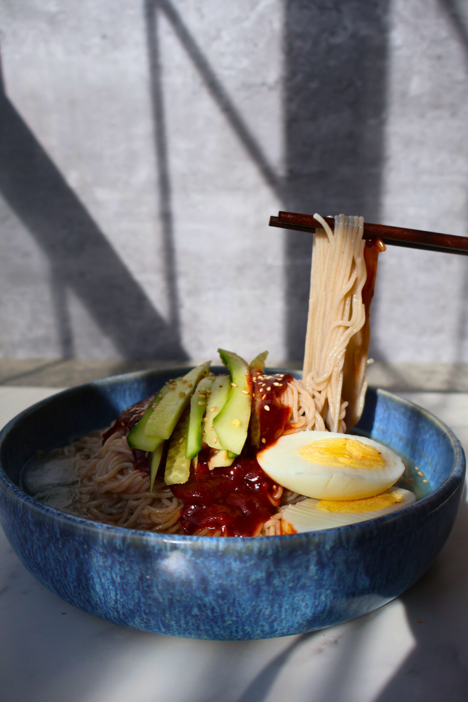 Korean bibim naengmyeon with noodles lifted by chopsticks, spicy gochujang sauce, cucumber, and boiled egg in a blue bowl