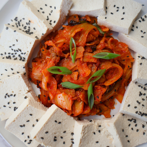 Overhead view of dubu kimchi on a white plate — thick slices of firm tofu arranged in a circle around a mound of spicy stir-fried kimchi and pork belly, garnished with sliced green onions and black sesame seeds, on a marble surface