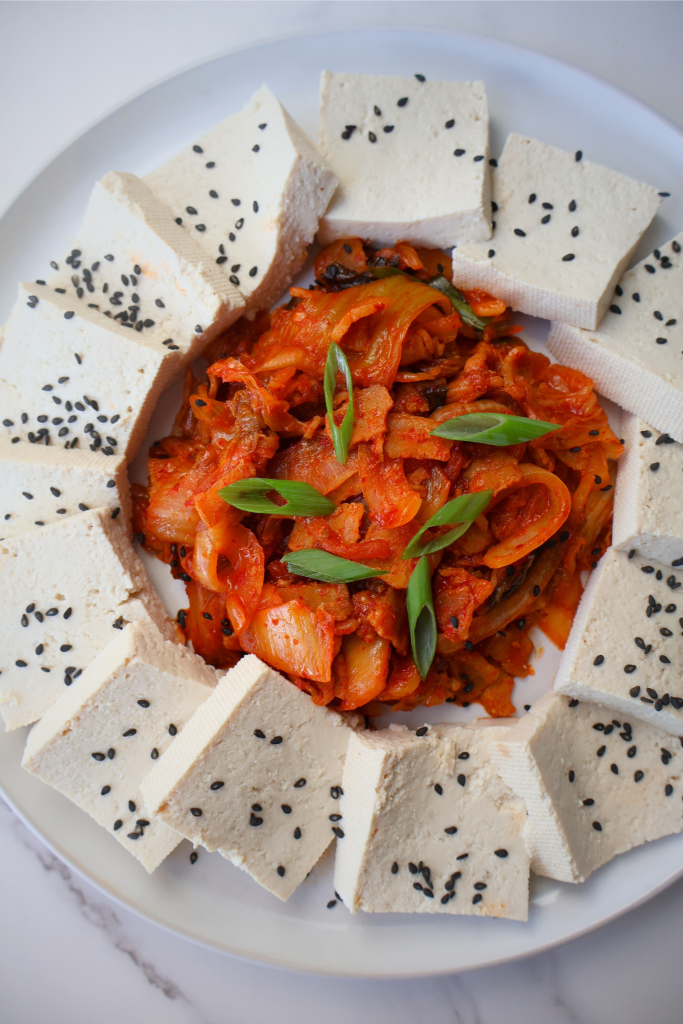 Overhead view of dubu kimchi on a white plate — thick slices of firm tofu arranged in a circle around a mound of spicy stir-fried kimchi and pork belly, garnished with sliced green onions and black sesame seeds, on a marble surface
