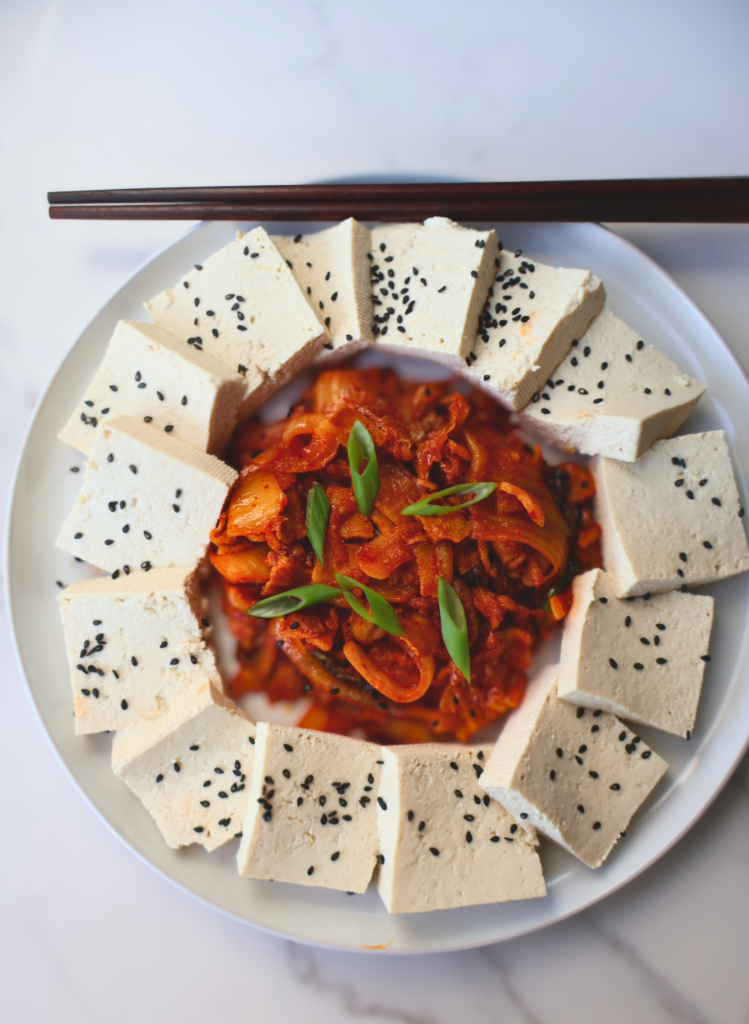 Overhead view of dubu kimchi on a white plate with wooden chopsticks resting on the rim — thick slices of firm tofu arranged in a ring around spicy stir-fried kimchi and pork belly, garnished with sliced green onions and black sesame seeds, on a white marble surface