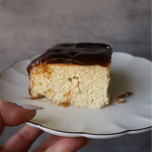 Slice of Balkan-style tres leches cake topped with caramel glaze on a white scalloped plate.