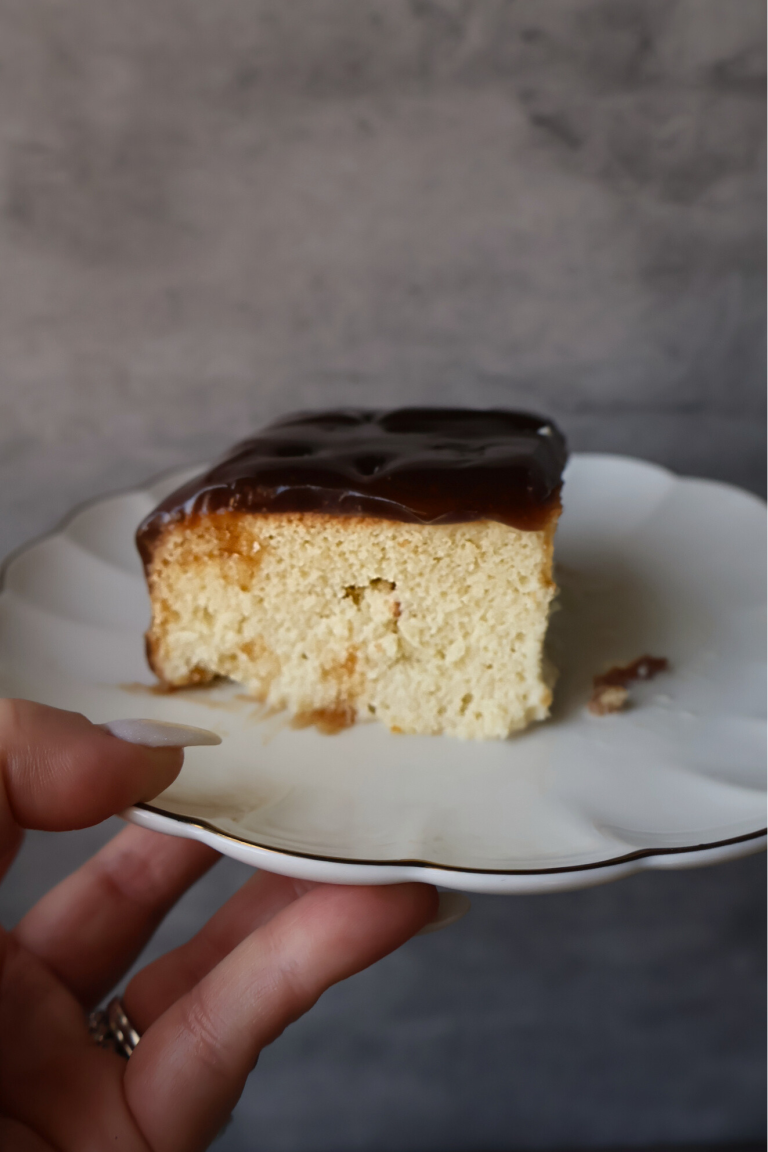 Slice of Balkan-style tres leches cake topped with caramel glaze on a white scalloped plate.