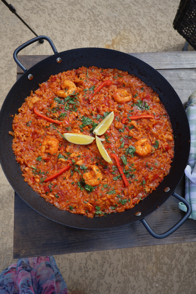 Full paella pan filled with tomato rice, chicken, and shrimp on a wooden surface outdoors, garnished with lemon wedges, parsley, and red bell pepper strips