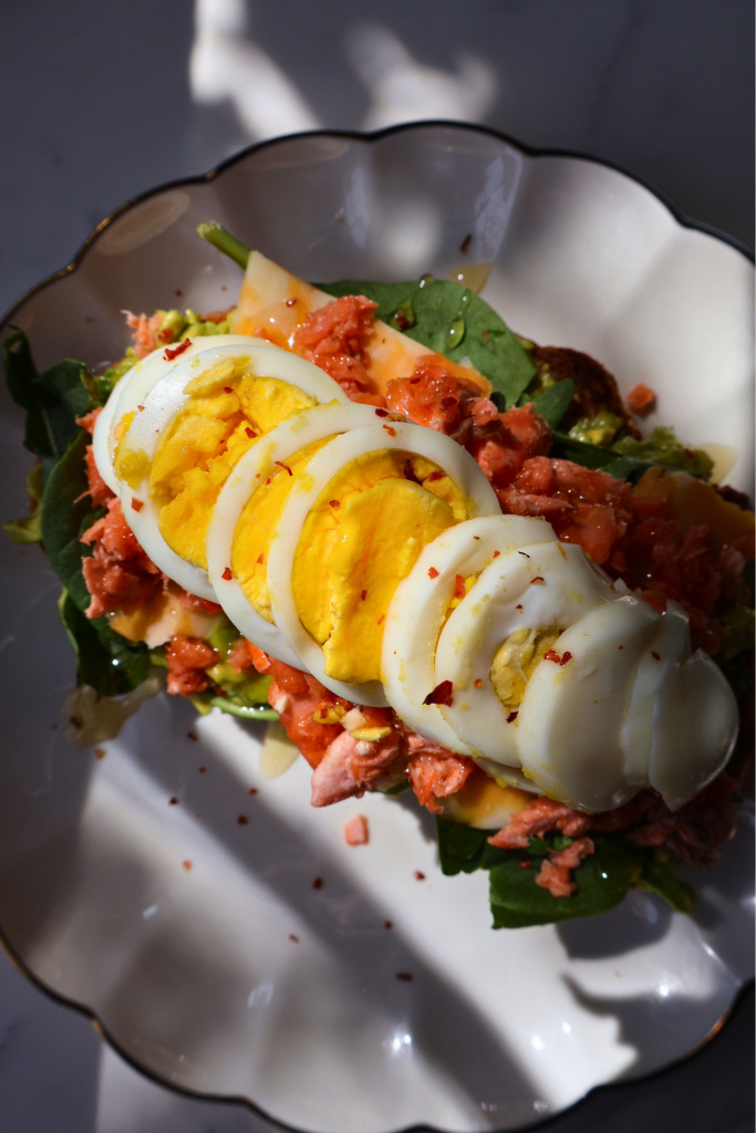 Angled side view of open faced salmon and avocado sandwich on sourdough on a white scalloped gold rimmed plate showing the height and layers of arugula, flaked salmon, and sliced boiled egg with natural light
