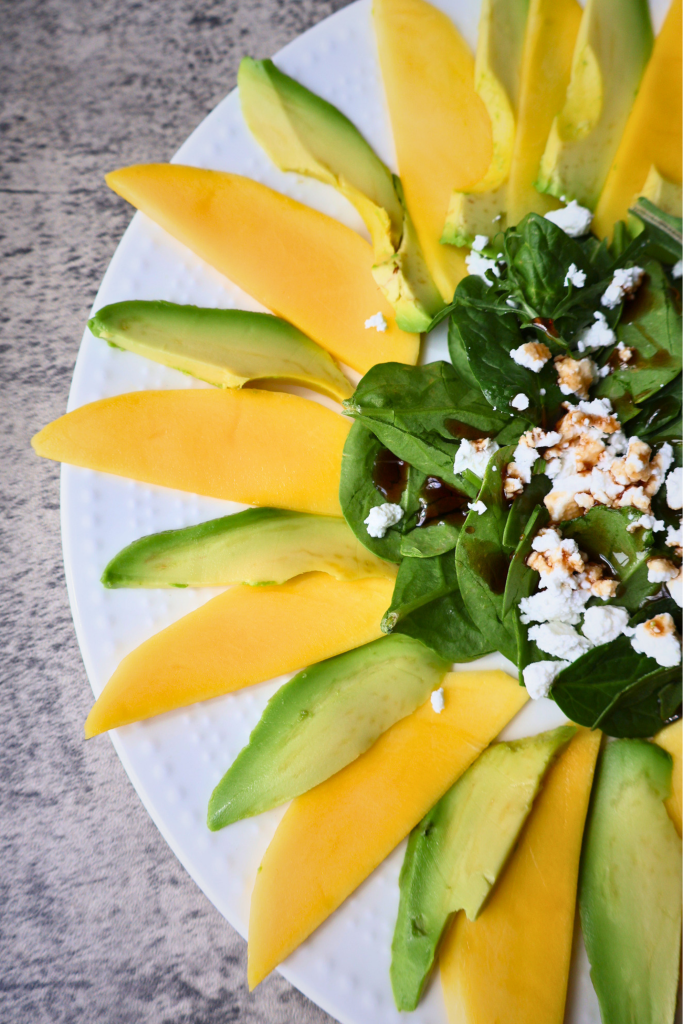 Mango and avocado carpaccio salad arranged in a sunflower pattern on a white round plate, topped with arugula, spinach, Bulgarian feta crumbles, chopped pistachios, and pomegranate molasses