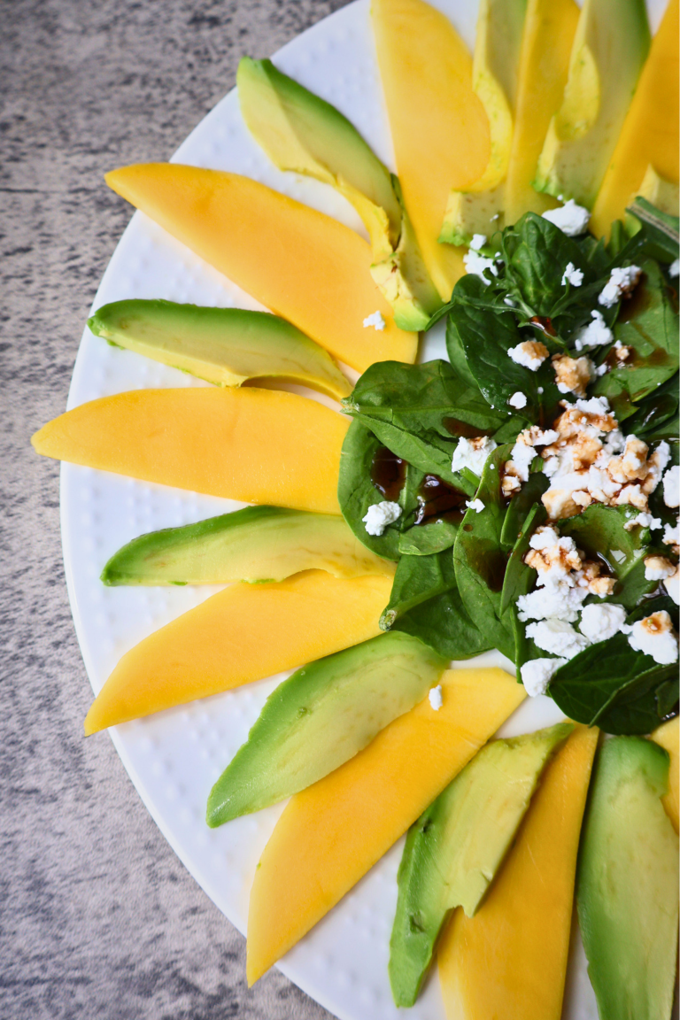 Mango and avocado carpaccio salad arranged in a sunflower pattern on a white round plate, topped with arugula, spinach, Bulgarian feta crumbles, chopped pistachios, and pomegranate molasses