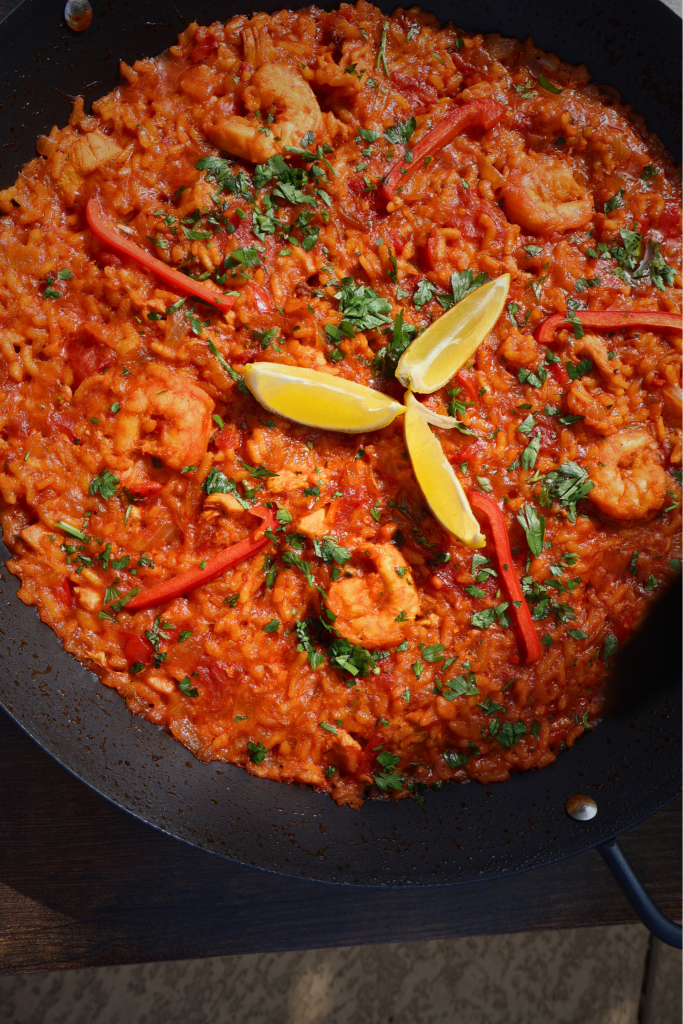 Overhead shot of tomato paella in a black paella pan showing the rich orange bomba rice with chicken, shrimp, red bell pepper, fresh parsley, and lemon wedges.