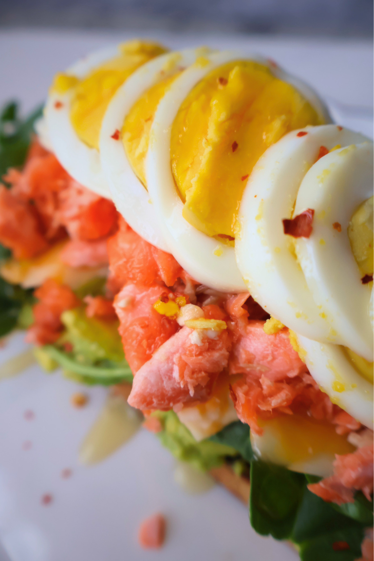 Close up of open faced salmon and avocado sandwich on sourdough showing fanned sliced boiled egg over flaked fresh salmon and arugula with crushed red pepper and honey drizzle