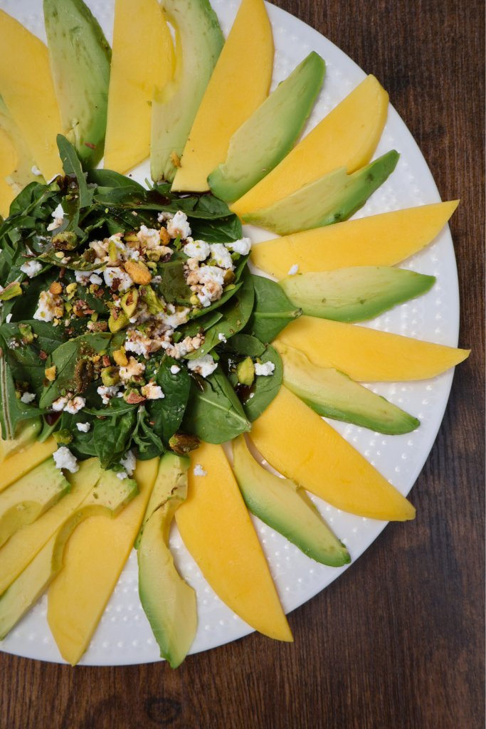 Mango and avocado carpaccio salad arranged in a sunflower pattern on a white round plate, topped with arugula, spinach, Bulgarian feta crumbles, chopped pistachios, and pomegranate molasses