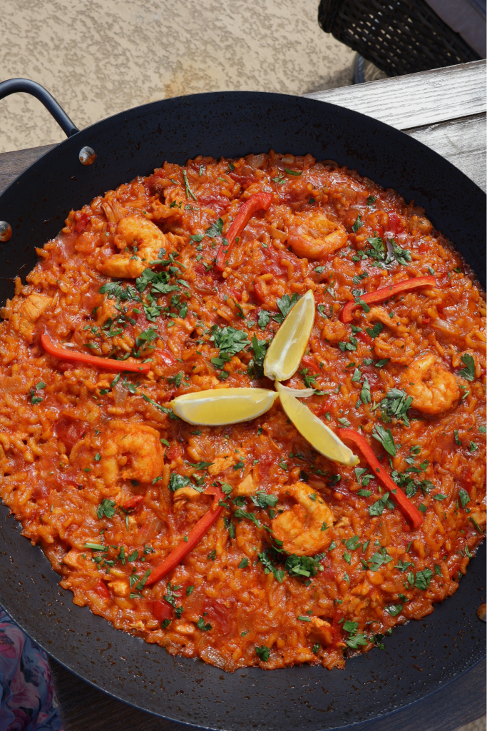 Tomato paella with chicken and shrimp in a large black paella pan, garnished with fresh parsley, lemon wedges, and red bell pepper strips, photographed from above