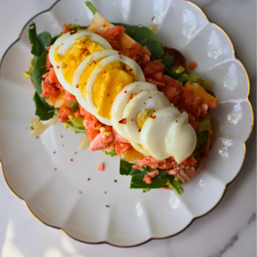 Close up shot of open faced salmon and avocado sandwich on toasted sourdough with sliced boiled egg fanned across flaked fresh salmon, arugula, crushed red pepper, and honey drizzle on a gold rimmed plate.