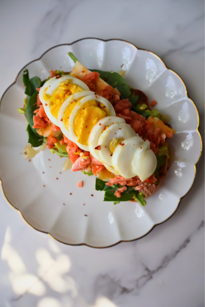 Close up shot of open faced salmon and avocado sandwich on toasted sourdough with sliced boiled egg fanned across flaked fresh salmon, arugula, crushed red pepper, and honey drizzle on a gold rimmed plate.