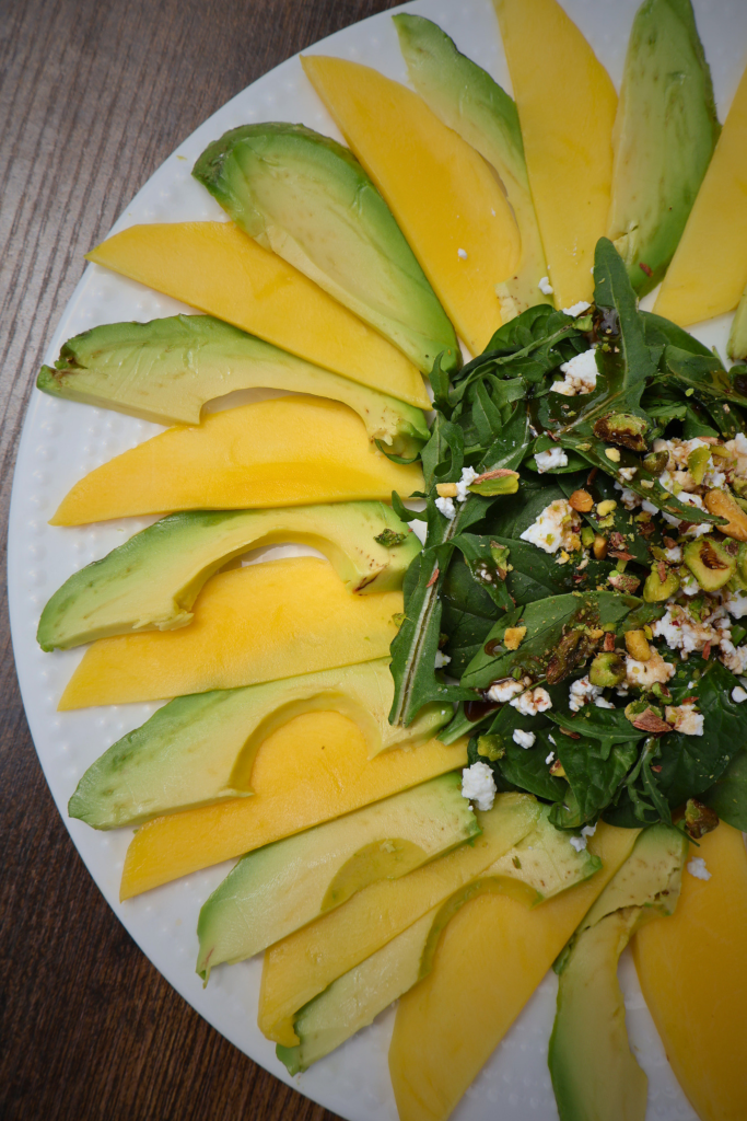 Close up of Mango and avocado carpaccio salad arranged in a sunflower pattern on a white round plate, topped with arugula, spinach, Bulgarian feta crumbles, chopped pistachios, and pomegranate molasses